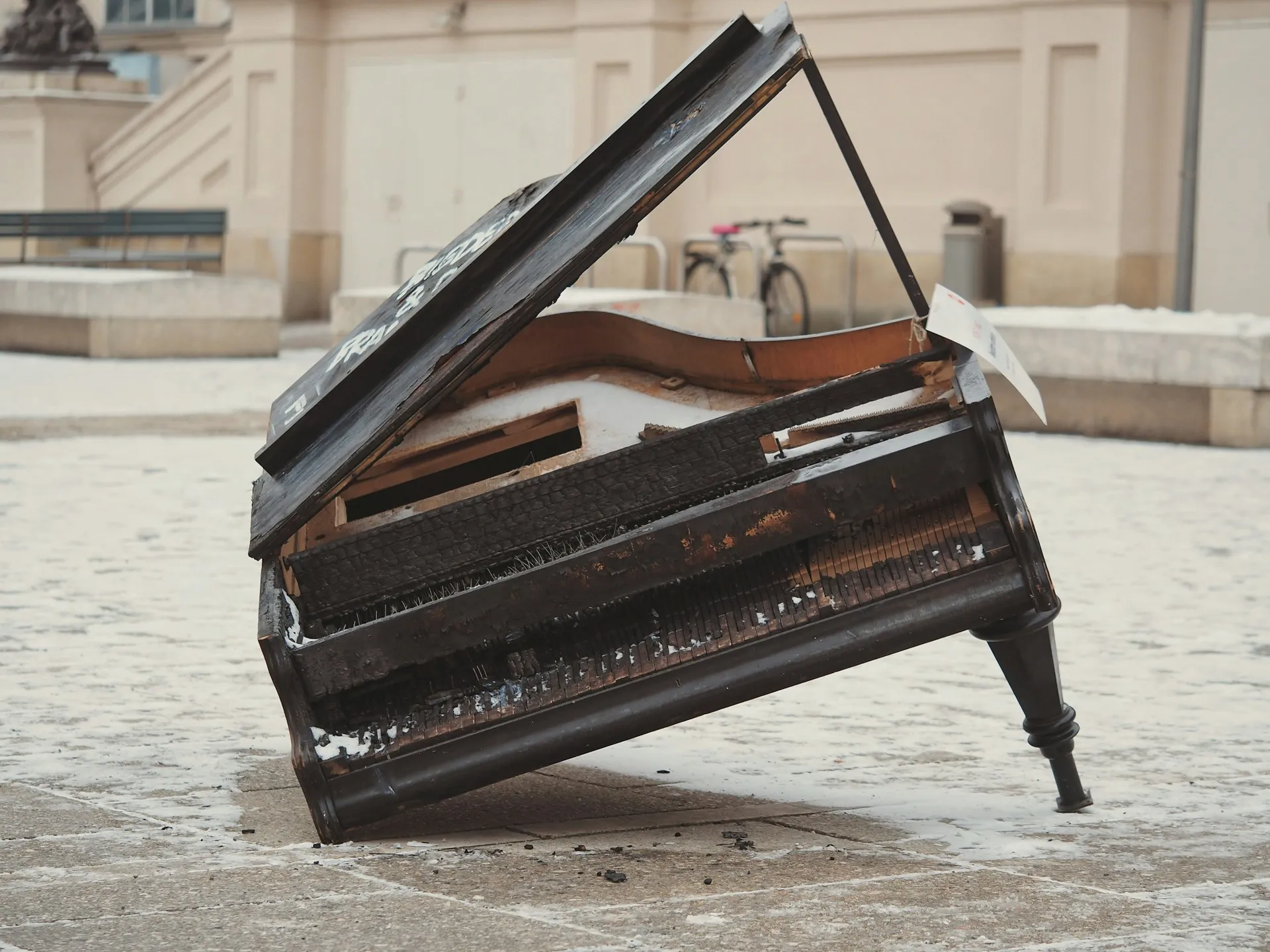 A black grand piano, missing a leg and reduced to a leaning husk.