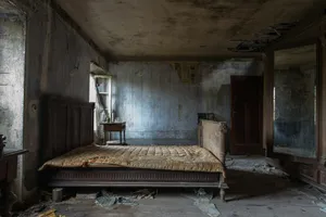 A large dilapidated room whose focal point is a bare bed frame, with a wooden dresser and nightstands around the outskirts.