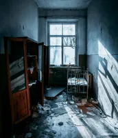 A narrow room featuring broken furniture and a floor littered with indecipherable pieces. The room is all the more striking due to the stark light pouring in through a broken window that occupies most of the back wall.